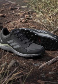 Chaussures de randonnée Adidas Terrex noires et grises avec semelles robustes placées sur un terrain rocheux et sec avec de l'herbe et des pierres dispersées.
