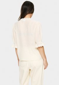 Cremfarbene, strukturierte Bluse mit puffärmeln und gewellten Spitzenakzenten an den Bündchen. Schlichte Rückseite. Kombiniert mit hellen Hosen.