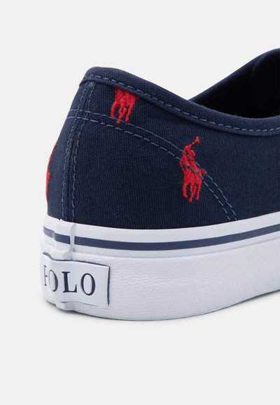 Sapatilha de lona azul-marinho com logotipo bordado em vermelho, sola de borracha branca e detalhe em risca azul-marinho. O remendo texturizado na parte de trás exibe o nome da marca.