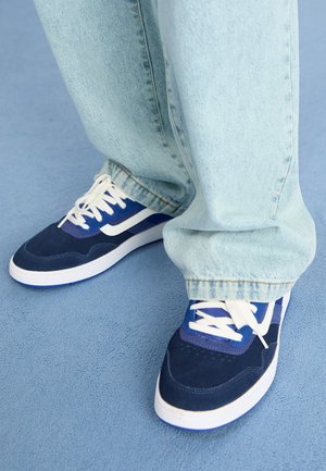 Pies con zapatillas deportivas azules y blancas y vaqueros azul claro, de pie sobre un suelo azul texturizado.