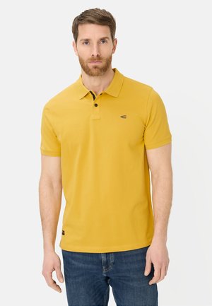 Mężczyzna o krótkich brązowych włosach i brodzie, ubrany w musztardowożółtą koszulkę polo i niebieskie dżinsy, stojący na tle jednolitego, jasnego tła.