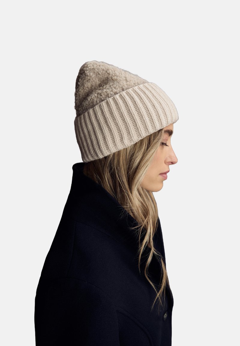 Berretto in maglia beige con parte superiore texturizzata e soffice. Presenta un bordo a costine ed è indossato con un cappotto nero. Vista di profilo.