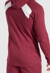 Sudadera marrón con acentos blancos en los hombros. Presenta un corte relajado, textura suave y capucha. La vista trasera muestra detalles de costura.