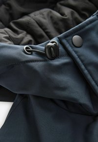 Tejido de chaqueta azul marino con textura suave. Presenta herrajes negros, un cordón ajustable y un forro visible en negro y gris oscuro.