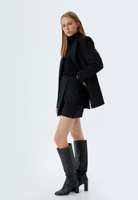 Zwarte blazer, geribbelde zwarte coltrui, zwarte mini-jurk en zwarte leren laarzen tot aan de knie met hakken. Gladde texturen, tailored ontwerp.