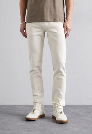 Homme portant un jean slim écru, des baskets décontractées beiges et une chemise marron à manches courtes, debout sur un sol gris.
