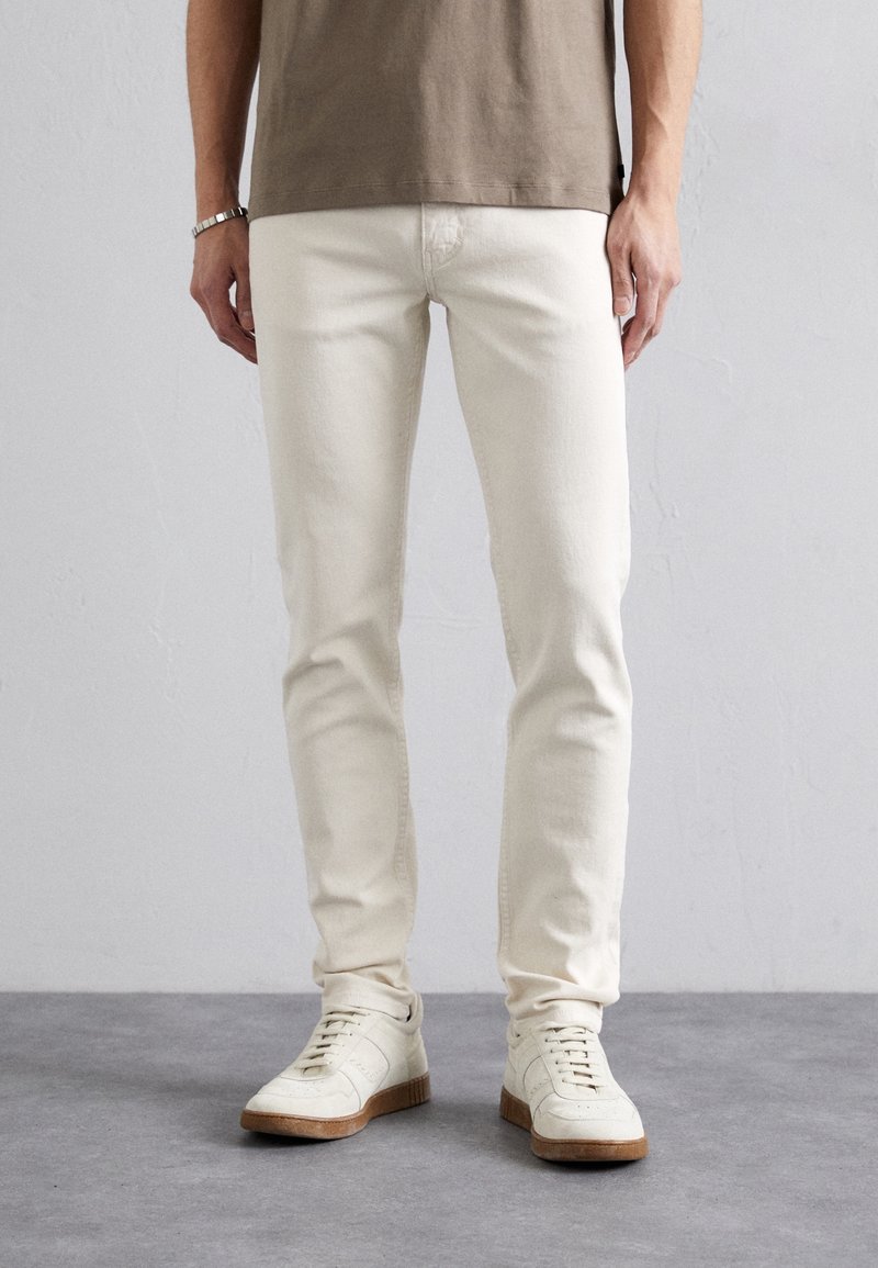 Homme portant un jean slim écru, des baskets décontractées beiges et une chemise marron à manches courtes, debout sur un sol gris.