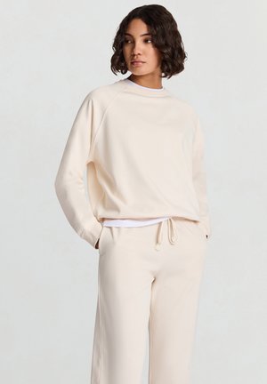 Hellbeiger Pullover mit lockerem Schnitt, Raglanärmeln und einem abgerundeten Ausschnitt, kombiniert mit passenden beigen Hose mit Kordelzug.