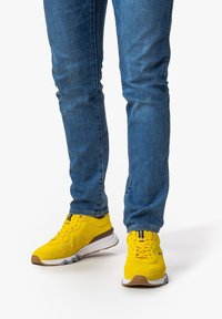 Gula sneakers med en strukturerad mockaövre, gummisula och kontrasterande grå detaljer, i kombination med tajta blå jeans.