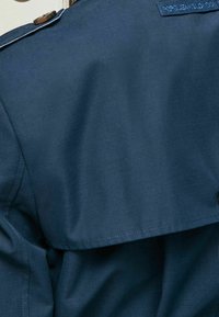 Tmavě modrá bunda s hladkou texturou, klopovým límcem a knoflíkovou aplikací na rameni. O značce je viditelná visačka na zadní straně.