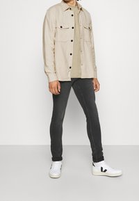 Beżowa koszula zapinana na guziki z dwoma kieszeniami na piersi, założona na oliwkową koszulkę. Noszona z czarnymi dopasowanymi dżinsami i białymi trampkami.