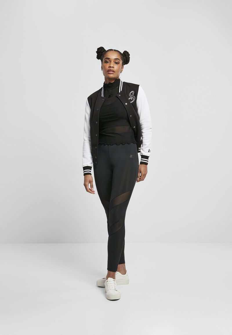 Čierno-biely varsity jacket prehozený cez čierny priliehavý top, doplnený s čiernymi legínami s priesvitnými panelmi. Biely tenisky dopĺňajú celkový vzhľad.
