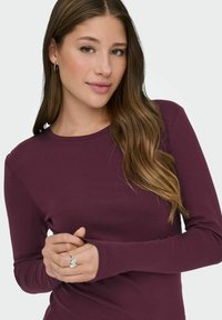 Burgunderfarbener Langarm-Oberteil aus weichem Stoff, mit Rundhalsausschnitt und enganliegenden Ärmeln. Die Hände sind gefaltet, wobei ein Ringdetail zur Geltung kommt.