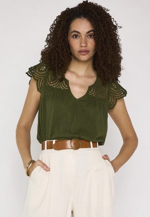 Donna con capelli ricci che indossa una maglia verde oliva con maniche in pizzo, pantaloni beige a vita alta e una cintura marrone con fibbia rotonda, mani nelle tasche.