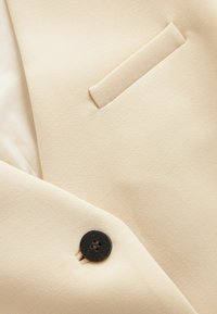 Gros plan sur un tissu beige avec un bouton noir et un détail de poche passepoilée horizontale.