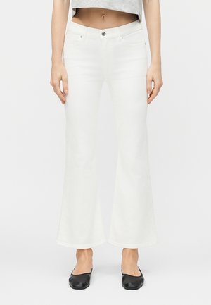 Γυναίκα που φοράει άσπλο τζιν ψηλόμεσο με φαρδιά μπατζάκια, γκρι κοντό τοπ και μαύρα επίπεδα παπούτσια, στέκεται μπροστά σε έναν απλό ανοιχτό φόντο.