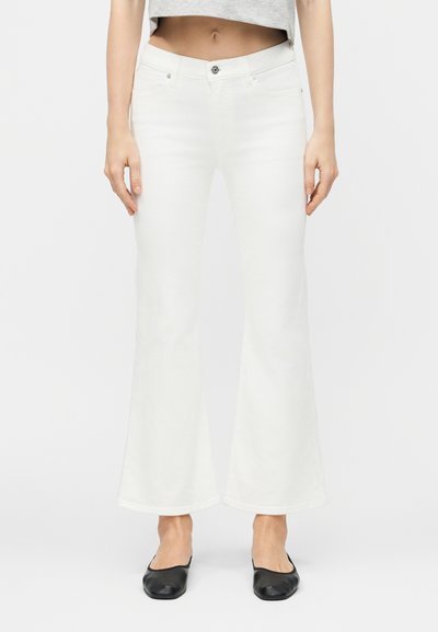 Femme portant un jean blanc évasé taille haute, un haut gris court, et des chaussures plates noires, debout devant un fond clair uni.