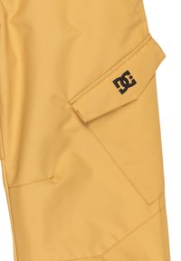 Gule vandtætte bukser med en glat tekstur, der har et sort logo-patch og vinklede designelementer på lommer og sømme.