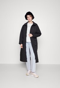 Long black quilted coat with a high collar, featuring side pockets, worn over a white shirt and light denim jeans, paired with sneakers.