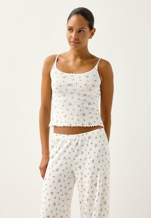 Kvinde iført hvidt ribbet blomsterprint camisole og matchende bukser med vide ben, stående mod en ensfarvet lys baggrund, kiggende til sin venstre side.