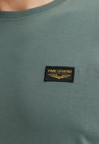 Donkergroene katoenen t-shirt met een ronde hals en een zwarte patch met gouden tekst en een vleugelontwerp met het label "PME LEGEND American Classic."