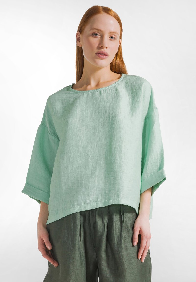 Vrouw met stijl rood haar, gekleed in een los lichtgroen linnen top met driekwart mouwen en donkergroene linnen broek, staand tegen een witte achtergrond.