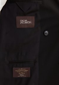Binnenkant van een zwarte blazer met een bruine label waarop "OSCAR JACOBSON" staat en een andere label die "VITALE BARBERIS CANONICO" aangeeft. Soepele stof.