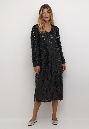 Schwarzes, langärmeliges Kleid mit Paillettenverzierung, mit V-Ausschnitt und Seitenschlitzen. Trägt silberne Ballerinas und minimale Accessoires.