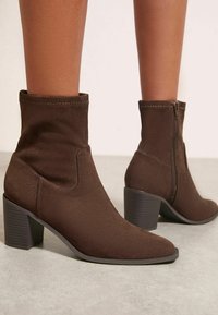 Brūnas zamša potītes ar norādītām pirkstiem un sakrautiem papēžiem. Iekļauj sānu rāvējslēdzēju un gludu tekstūru. Papēžiem ir horizontālas rievas.