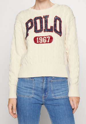 Femme portant un pull crème en maille torsadée avec "POLO 1967" en rouge et marine, associé à un jean bleu taille haute avec poches plaquées devant.