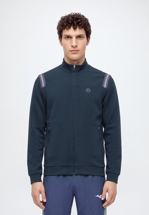 Homme aux cheveux bouclés portant une veste zippée foncée avec deux bandes réfléchissantes sur chaque épaule et un pantalon de sport bleu marine.