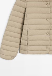 Giacca imbottita trapuntata color beige con cerniera e bottoni a scatto, tasca laterale e tessuto morbido e strutturato. La manica ha un bordo arrotondato.