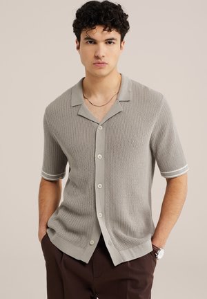 Noormees lühikeste käistega helehallist kootud nööpidega särgis ja tumedates pruunides pükstes, käed taskutes, seismas.