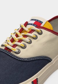 Καμβάς αθλητικό παπούτσι με ναυτικό μπλε, κρεμ, κόκκινες και κίτρινες λεπτομέρειες, λευκές και κίτρινες κορδόνια και εμφανές λογότυπο Tommy Jeans στη γλώσσα και στη σόλα.