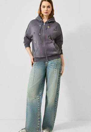 Femme portant un sweat à capuche gris zippé avec des ornements et un jean bleu délavé à jambes larges, debout devant un fond clair uni.