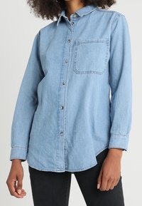 Ljusblå denimskjorta med långa ärmar, knappstängning fram och en enda bröstficka. Rundad fåll och avslappnad passform. Texturerat tyg.