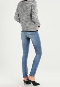 Personne portant un pull à motifs, un jean skinny bleu et des mules noires à talons aiguilles pointues, debout avec les mains dans les poches, dos face au spectateur.