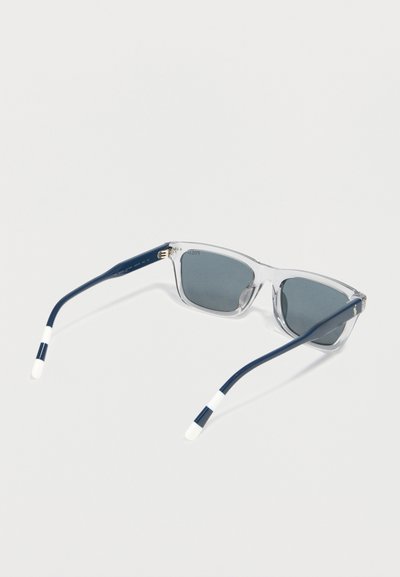 Lunettes de soleil à monture transparente avec verres foncés et branches bleu marine avec embouts blancs, posées ouvertes sur un fond clair.