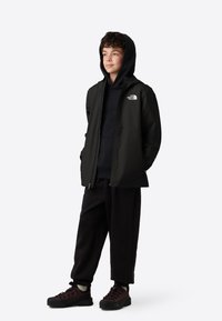 Černá lehká bunda s kapucí, na levé straně hrudi s logem, v kombinaci s tmavou mikinou s kapucí, černými kalhotami a odolnými botami.