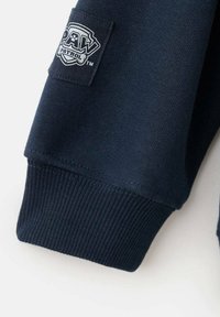 Manche de sweat-shirt bleu marine avec un poignet côtelé texturé et un patch en tissu portant le logo « PAW PATROL » en blanc et noir.