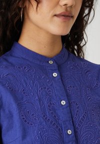 Camicia viola con bottoni a pressione e un intricato design paisley ricamato. Presenta un colletto alla mandarino e piccoli dettagli con bottoni bianchi.