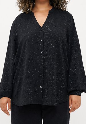 Zwarte blouse met lange mouwen en knoopsluiting, bedrukt met kleine witte spikkels, gedragen door een persoon met krullend haar, zichtbaar van nek tot heupen.