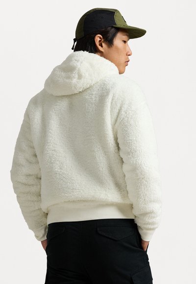 Hvit, fluffy hettejakke i fleece med ribbestrikket kant, brukt over mørke bukser. Modellen har en grønn caps som delvis dekker mørkt hår.