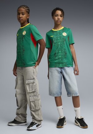 Zwei Jugendliche stehen nebeneinander und tragen grüne Senegal-Fußballtrikots mit gelben Kragen, lässige Shorts und Cargo-Hosen sowie schwarze Turnschuhe.