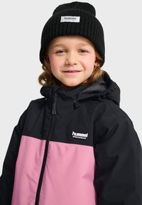 Veste à capuche noire et rose avec une fermeture éclair à l'avant, arborant un logo sur la poitrine et un bonnet noir côtelé sur la tête.