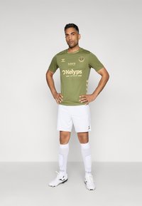 Grøn kortærmet fodboldtrøje med guld accenter, hvide shorts og knæhøje hvide sokker med grip-mønstre. Sorte fodboldstøvler.