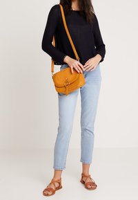 Borsa a tracolla gialla in suede con pattina, forma rettangolare e dettaglio in metallo circolare. Indossata con jeans blu chiaro e sandali marroni.