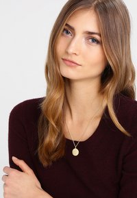 Goldene Anhänger-Halskette an einer zarten Kette, getragen von einem Model mit welligem, hellbraunem Haar, das einen dunkelbordeauxfarbenen Pullover trägt.