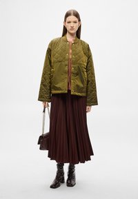 Olíva zöld steppelt bomberdzseki patentos gombokkal, barna pliszírozott szoknyával párosítva. A modell láncos pántú táskát tart, és fényes csizmát visel.