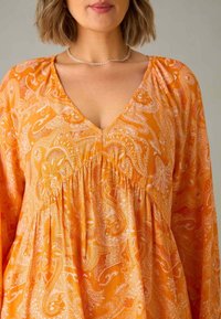 Vestido de manga larga con escote en V en brillante tela naranja que presenta un patrón de paisley. El detalle de la cintura fruncida añade textura.